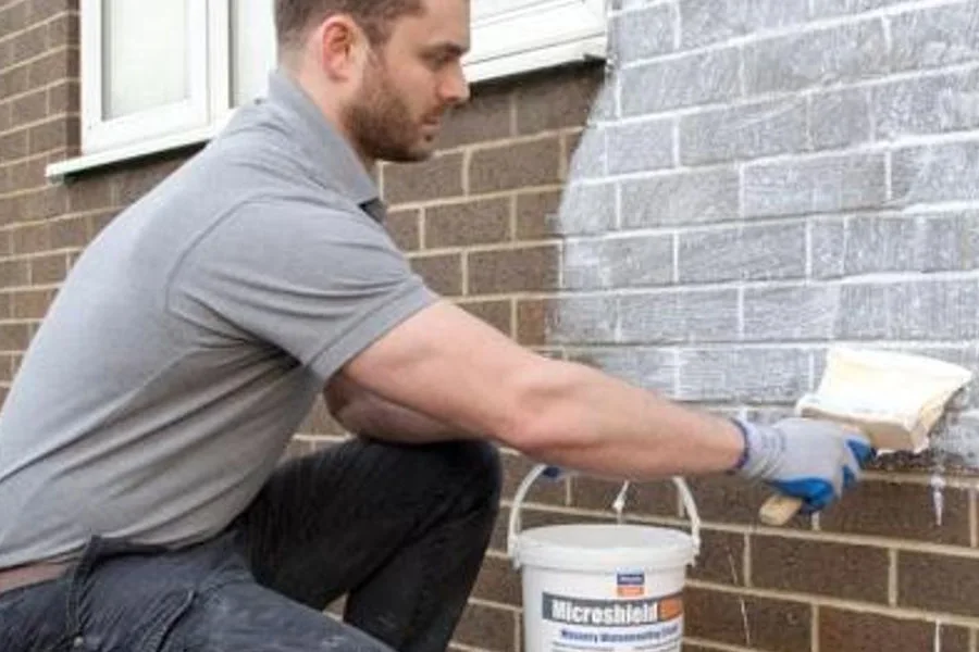 A worker brushing breathable waterproofing sealer onto a brick masonry wall